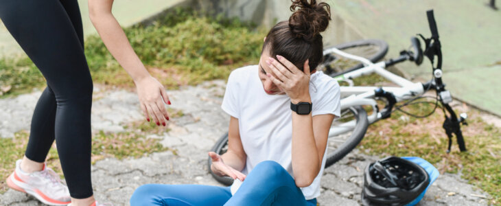 Crying Female Cyclist Sitting on Ground in Pain After Biking Accident While Female Approaching to Help Her